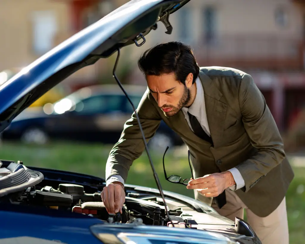 A man in a suit inspecting the engine of a car on the side of the road, looking focused as he holds his glasses in his left hand.