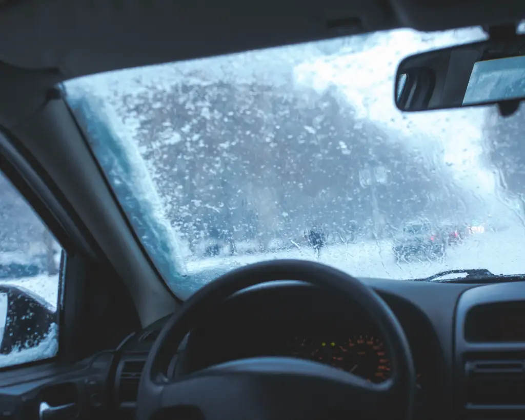 View from inside a car with a snow-covered windshield on a cold winter day, showing icy buildup and blurred visibility.
