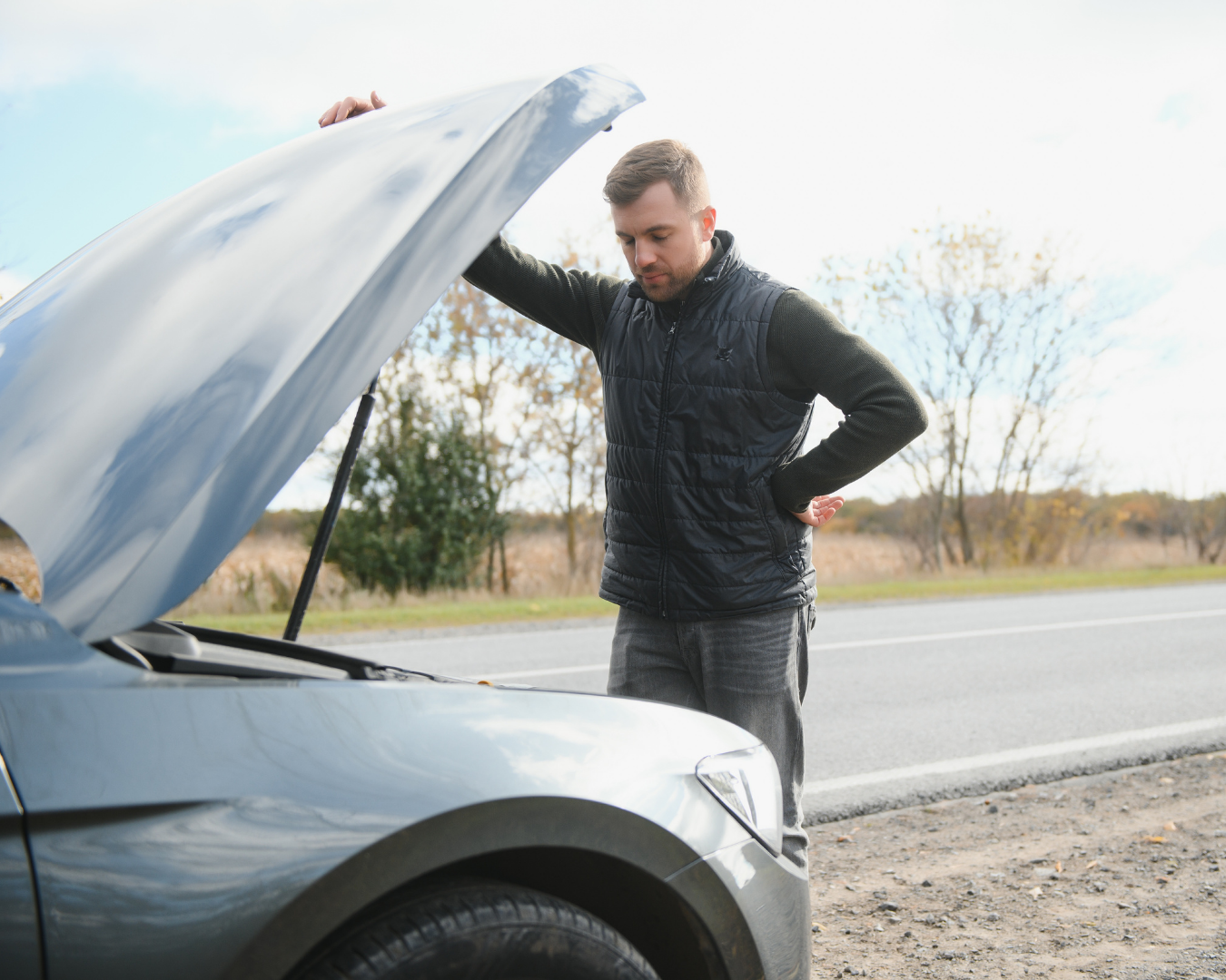 Man standing beside his broken-down car looking confused while checking the engine on the side of the road.